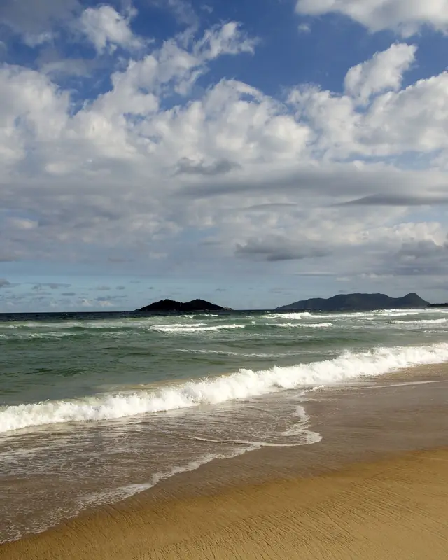 Praia do Campeche, em Florianópolis, com ondas quebrando e a Ilha do Campeche no horizonte
